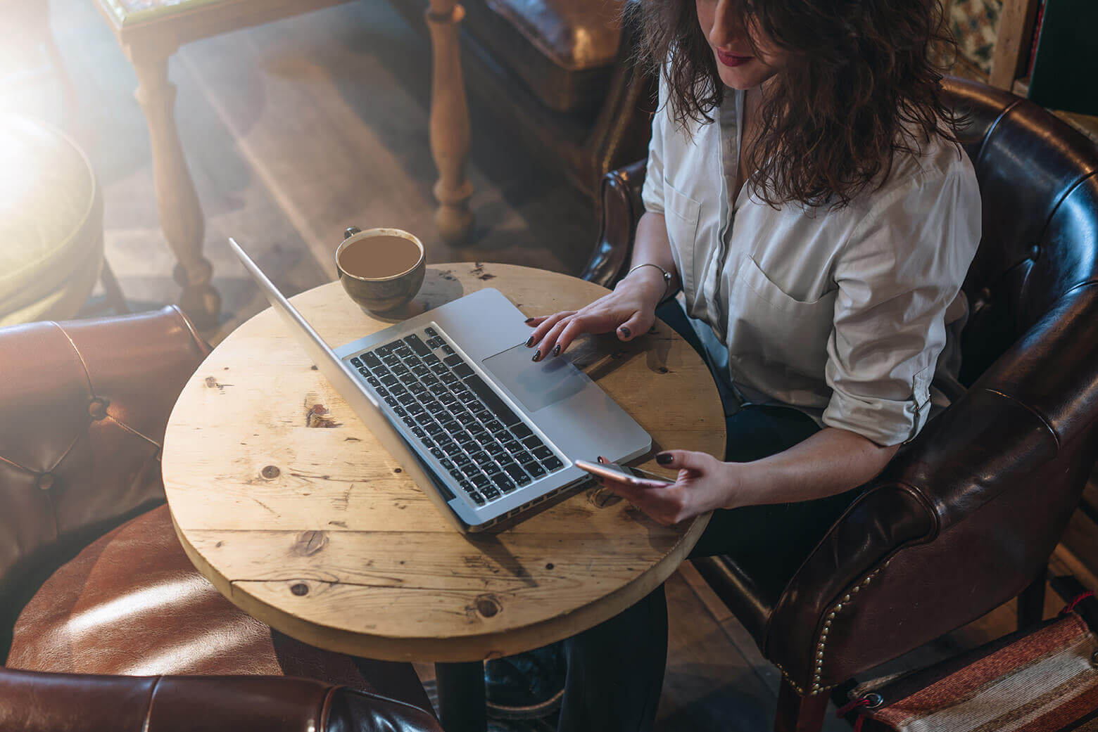 Woman sitting at a small round table looking at her phone and working on her laptop